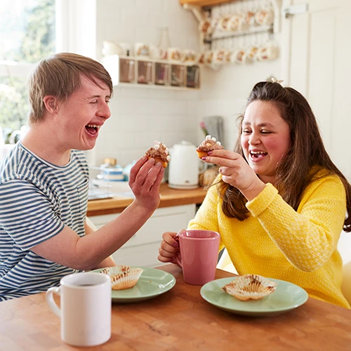 Young Downs Syndrome Couple Enjoying Tea And Cake In Kitchen At Home 
